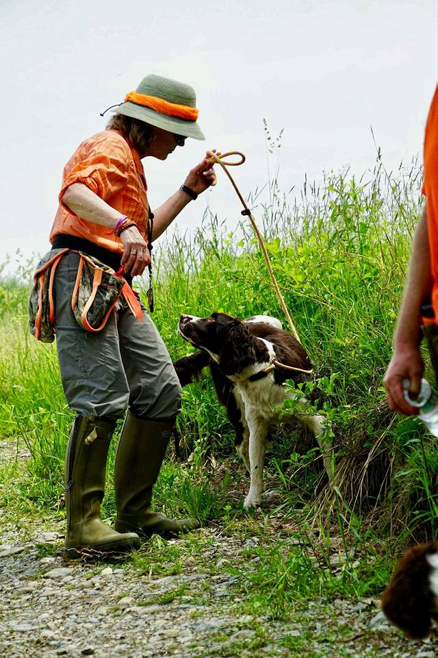 A woman in outdoor clothing and a hat interacts with a dog using a stick on a grassy trail, with another person partially visible on the right.
