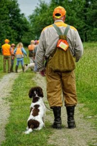 A man in hunting attire with an orange cap and a dog sits by his side, watching a group of people walking ahead in a grassy field.