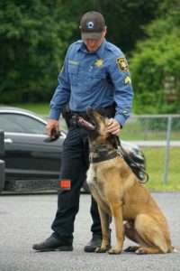 A police officer in uniform, with a badge and name tag visible, patting a german shepherd police dog beside a patrol car.