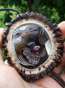 A detailed miniature painting of a dog with a bird in its mouth, encased in a round, textured frame, held by a hand against a green leafy background.