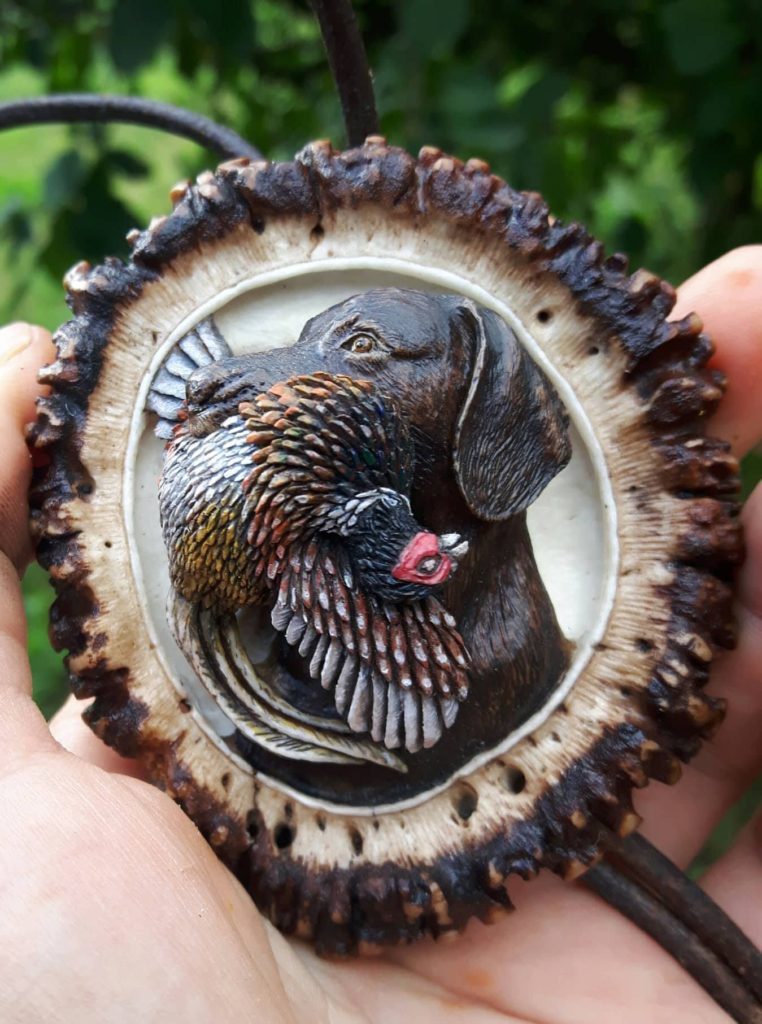 A detailed miniature painting of a dog with a bird in its mouth, encased in a round, textured frame, held by a hand against a green leafy background.