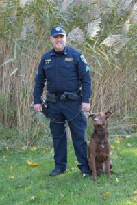 A police officer in uniform standing outdoors with a leashed brown dog, both posing in front of tall grass.
