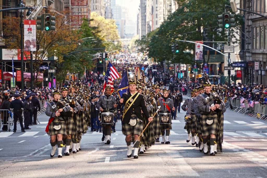 A procession of bagpipers in traditional scottish attire marching down a busy urban street during a parade, with onlookers lining the sidewalks.