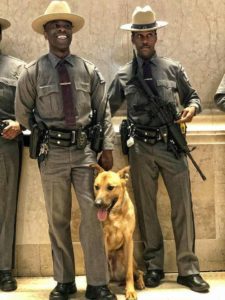 Two state troopers and a police dog standing in a building lobby, wearing uniforms and equipped with duty belts and firearms.