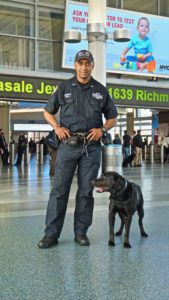 A police officer in uniform and his black dog stand in an airport terminal, under a sign with flight information displayed.