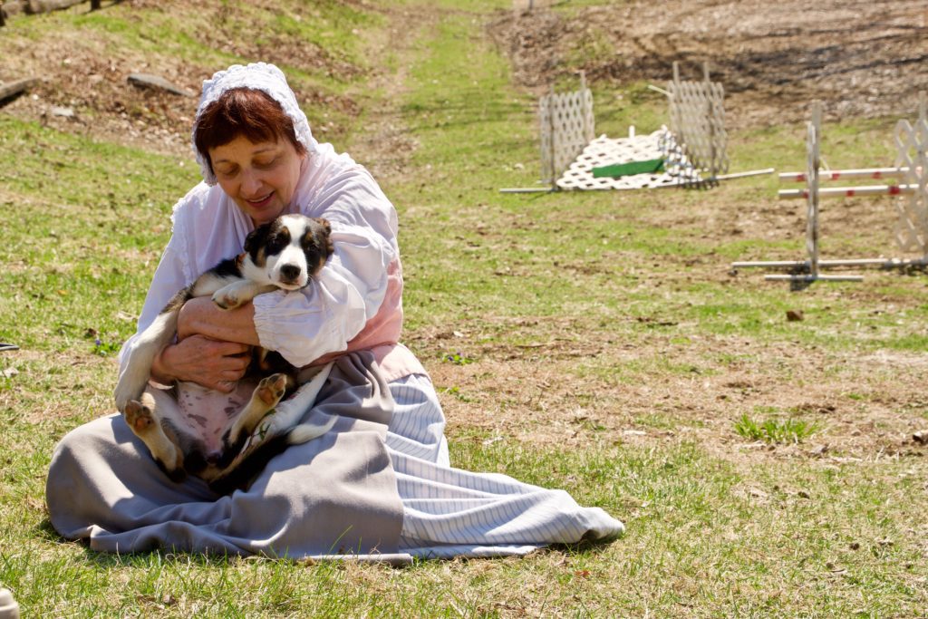 A woman in historical dress sitting on grass and holding a small black and white dog, with a fence and barren trees in the background.