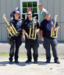Three police officers smiling and holding large trophies outside a building on a sunny day.