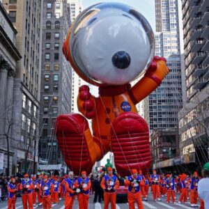 Big float of Snoopy wearing a red and orange astronaut suit being pulled by hundreds of people in orange and blue uniforms