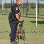 A police officer in uniform stands with a leashed patrol dog on a grassy field at the uspca national patrol dog trials in foley, alabama, 2023.