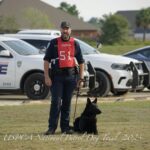 A police officer with a dog at the uspca national patrol dog trial in foley, alabama, 2023, with police cars in the background.
