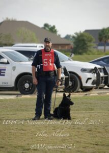 A police officer with a dog at the uspca national patrol dog trial in foley, alabama, 2023, with police cars in the background.