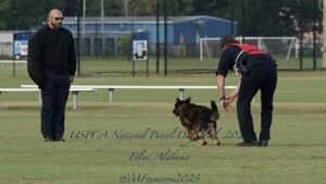 Two men and a german shepherd at a dog training competition in a grassy field, with text indicating it is the uspca national patrol dog trials in foley, alabama.